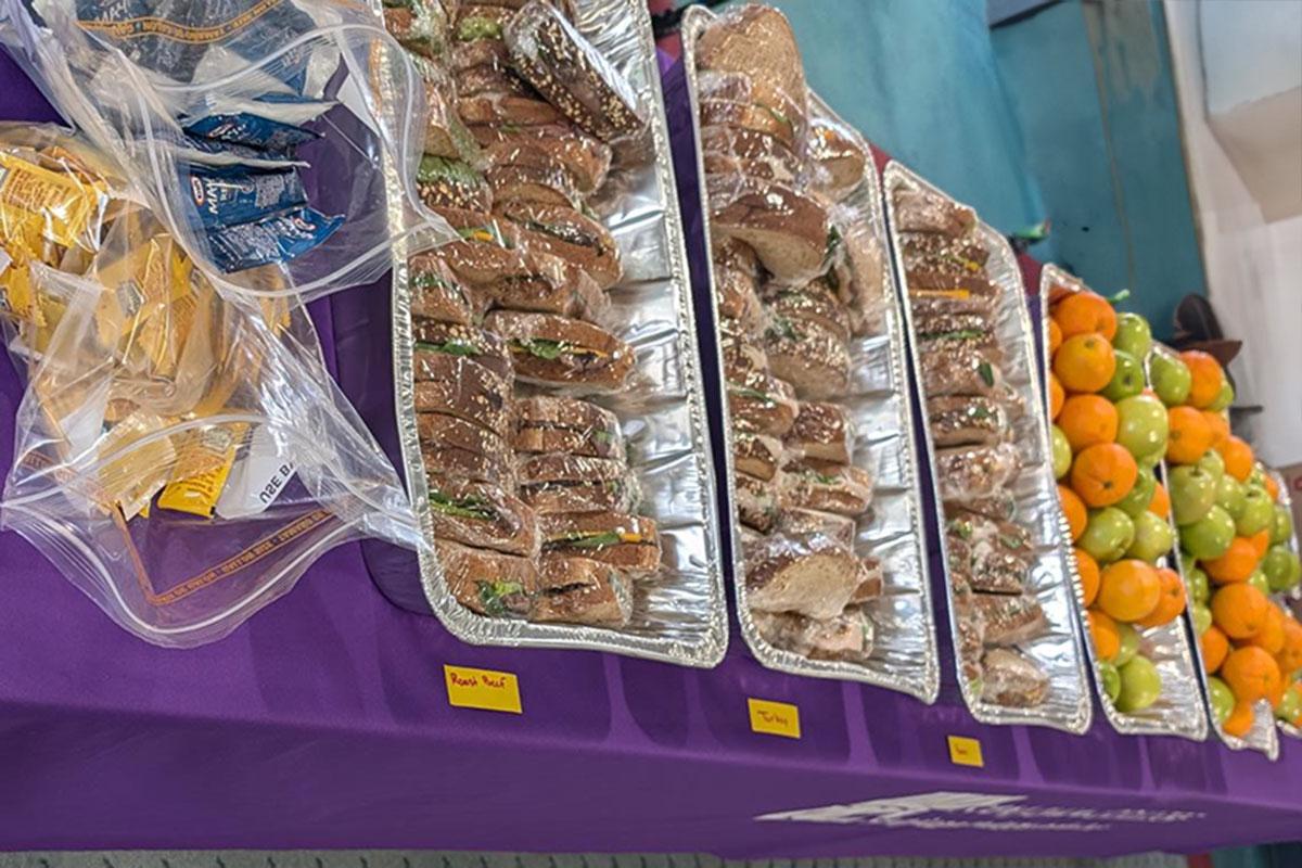 A purple table with variety of sauce packets, trays of sandwiches, and trays of oranges and green apples.